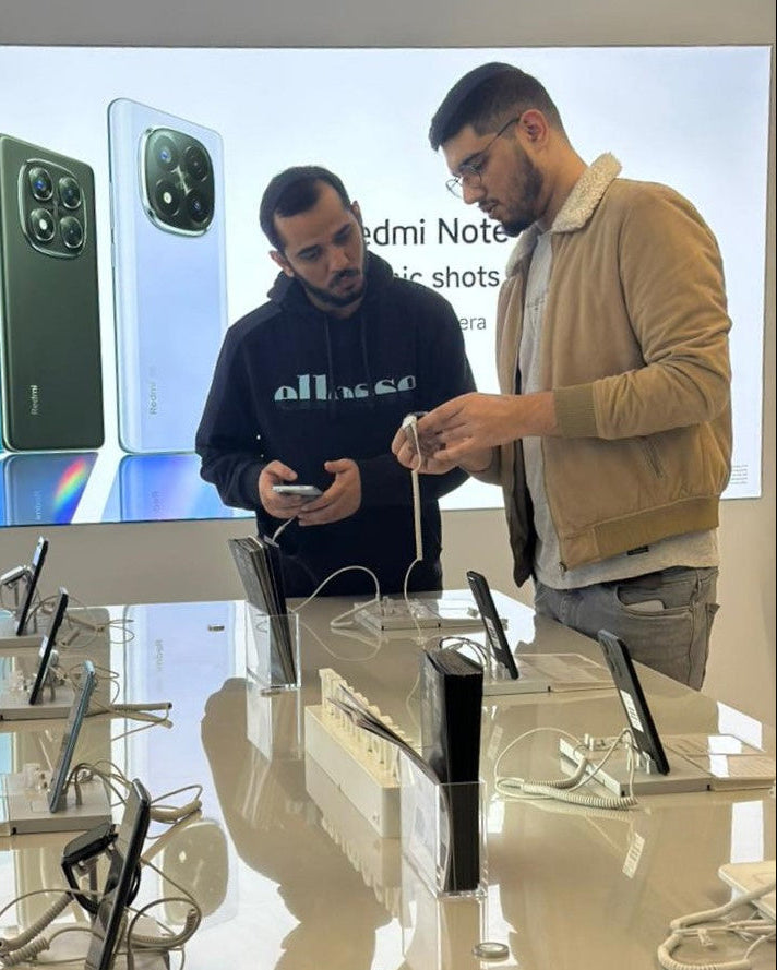 Two men looking at electronic devices on a display table with a screen showing phone models in the background.
